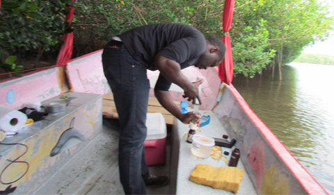 Abioudun Emmanuel Ayo-Bali, a geological sciences graduate student from Nigeria transfers a water sample from a syringe into a collection bottle while standing in a metal boat. He is surrounded by various research supplies and is facing away from the camera.